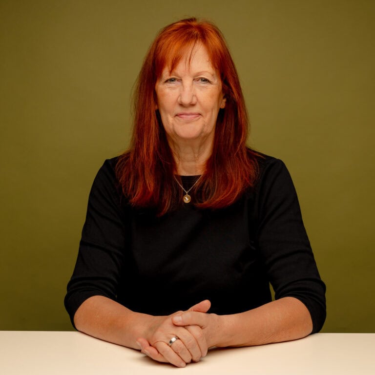 Frau sitzt an einem Tisch, Hände auf dem Tisch, Blick in die Kamera.